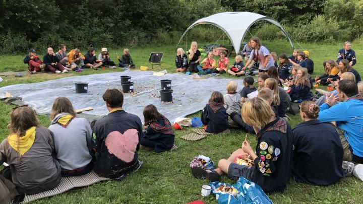 Spejdere samlet om fællesspisning på sommerlejren. Spejdere i alle aldre, der sidder omkring en pressening og spiser bl.a. revelsben til aftensmad. På presseningen står plastspande med pointangivelse på. Spejderne kan optjene point ved at ramme spandene med deres afgnavede revelsbensknogler